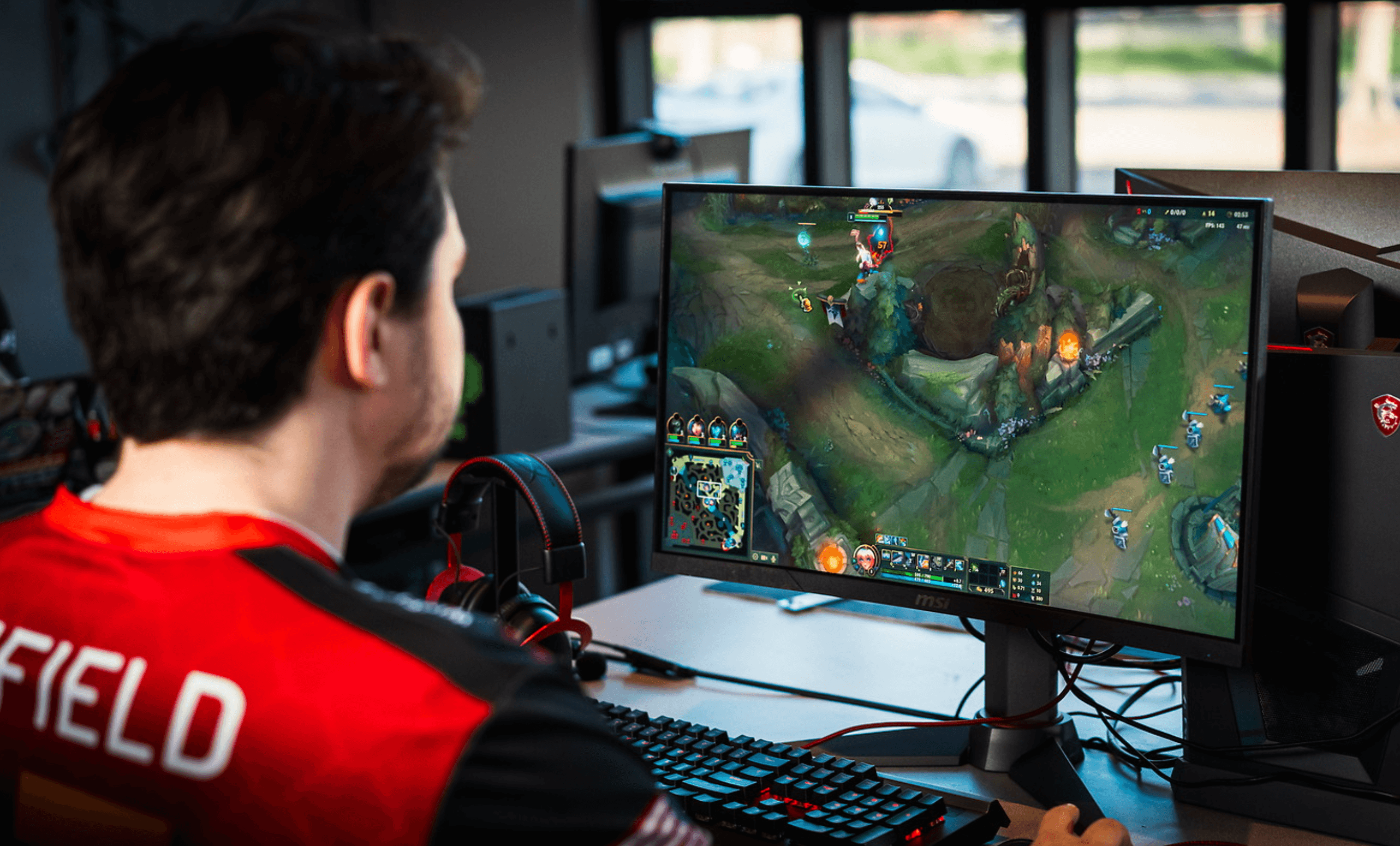 A person wearing a red jersey focuses on playing a strategy video game displayed on a computer monitor, with gaming accessories like a keyboard and headphones nearby; the room appears to have a modern setup with daylight streaming through large windows.