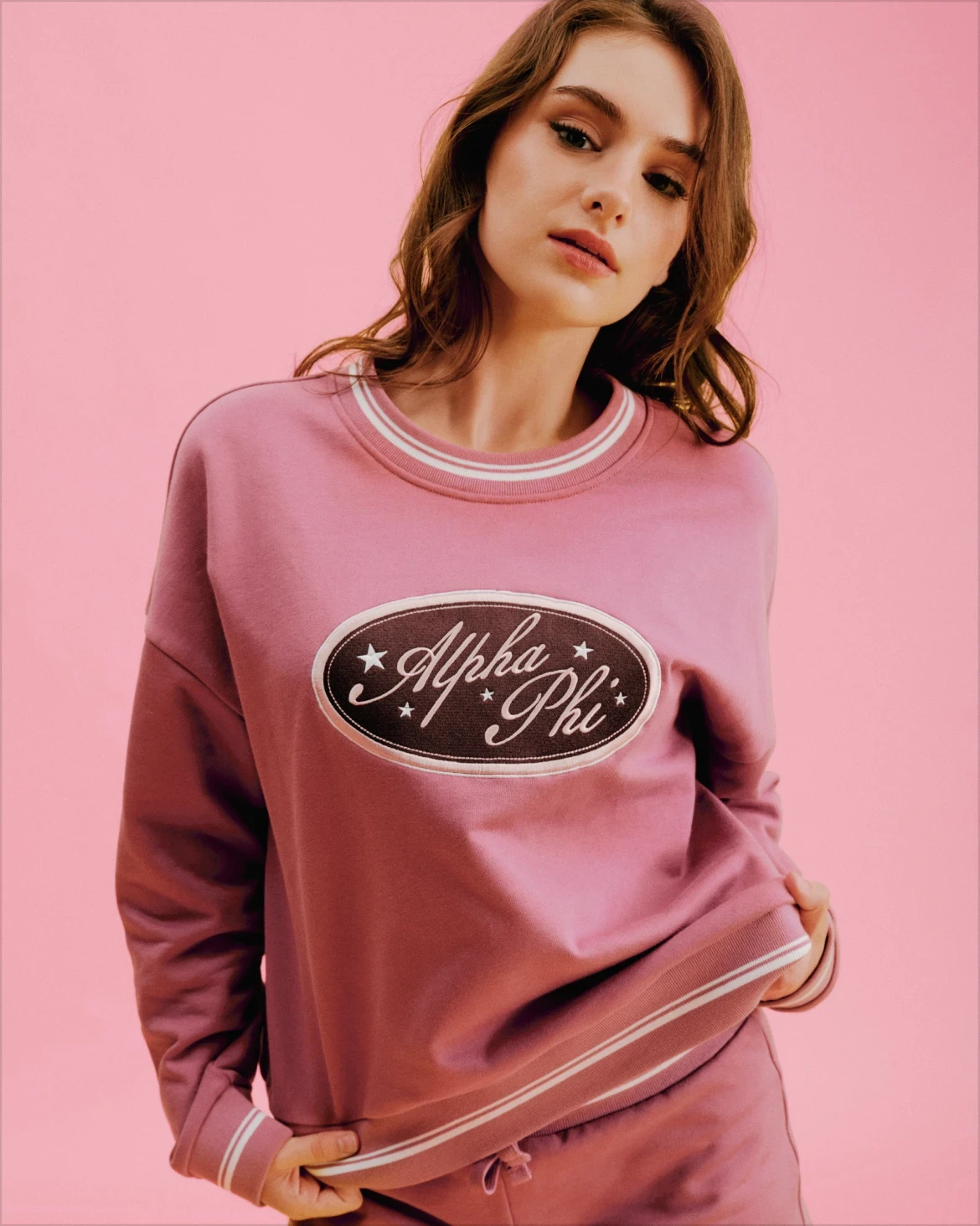 Model wearing a mauve Alpha Phi sweatshirt and matching shorts against a pink background.