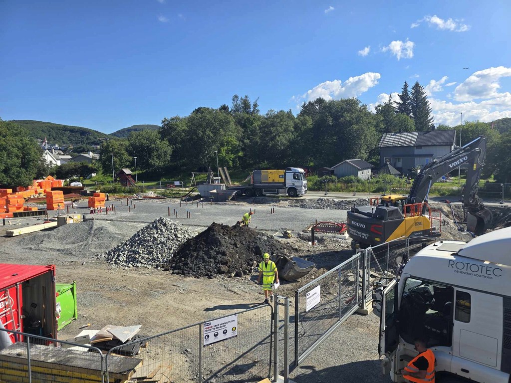 Byggeplass med byggegjerne, hauger med ulike masser, gravemaskiner og arbeidere i gule vester.