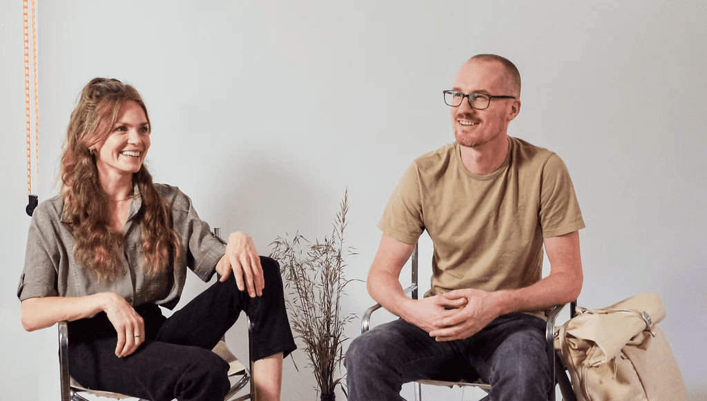 Two people are sitting on chairs indoors, smiling and engaged in conversation, with a minimalist backdrop featuring a small plant between them.