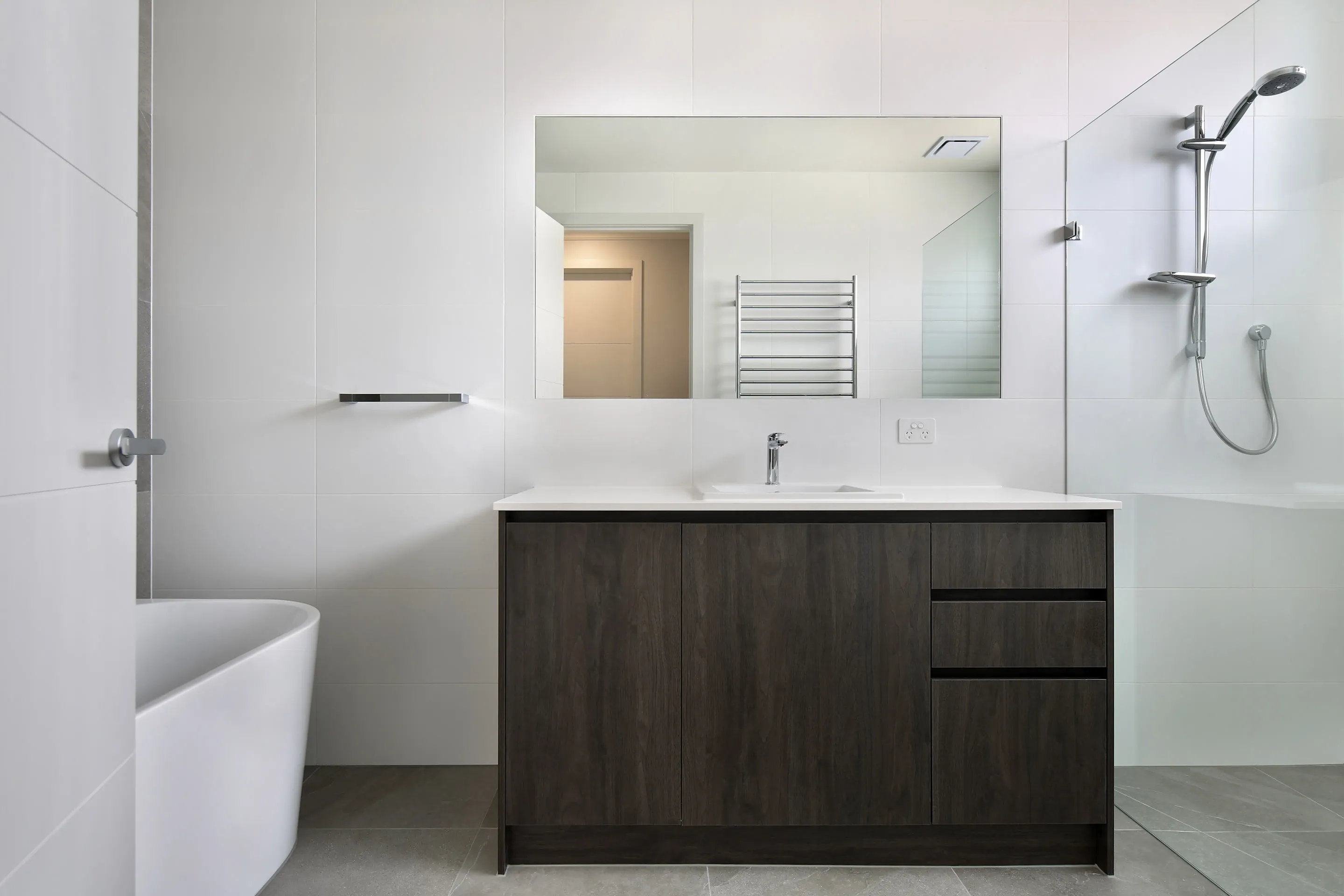 Bathroom with white tiles, a dark timber vanity with white stone top, freestanding bath, large mirror and a chrome rail shower