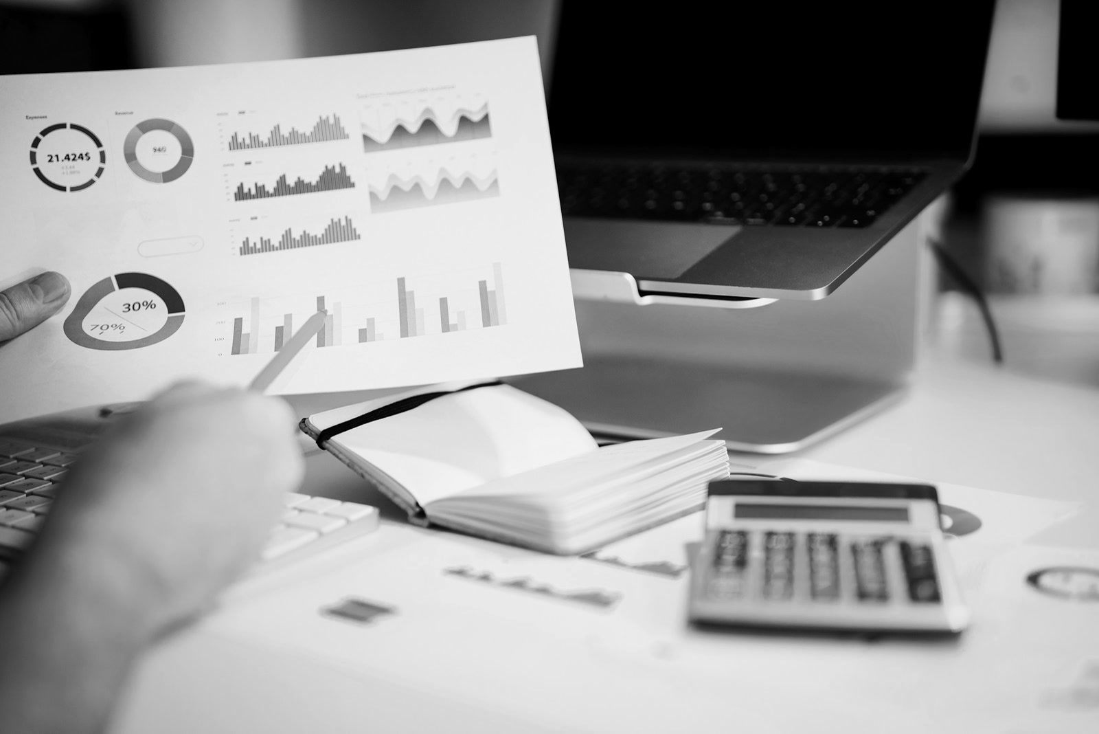 A hand with a pen over printed charts, a notebook and a calculator on a desk.