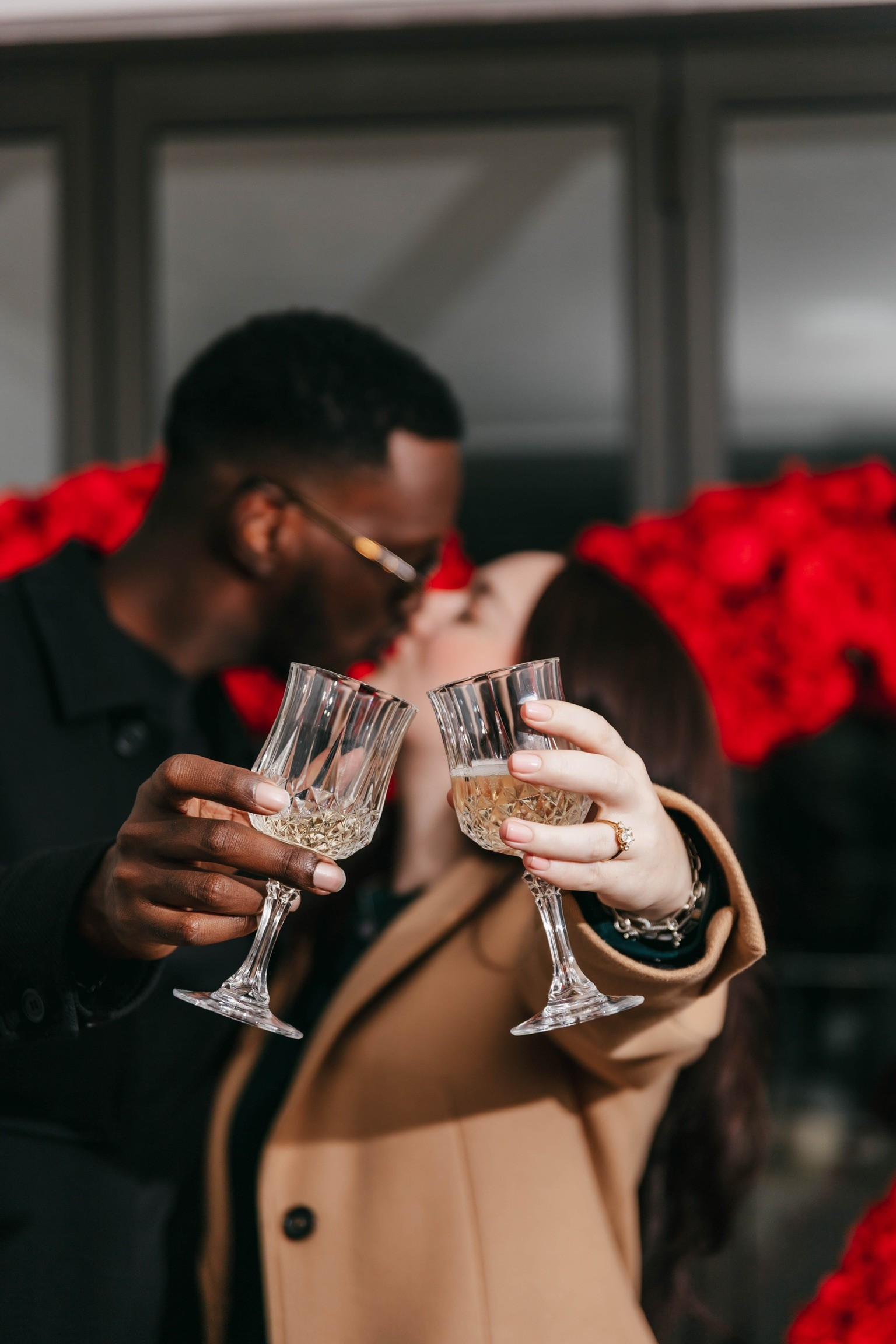 A bride and groom hold hands tenderly, the bride's bouquet of dried flowers hanging low. They stand in a softly lit, elegant interior, conveying romance.
