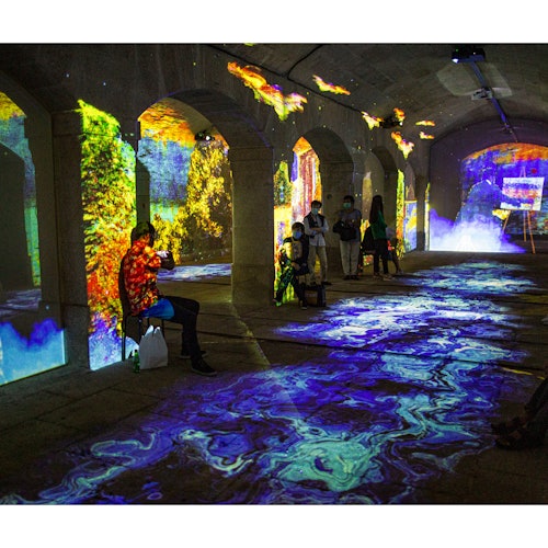 Pessoas em uma sala em arco com projeções digitais coloridas nas paredes e no chão, criando um ambiente vibrante e imersivo.