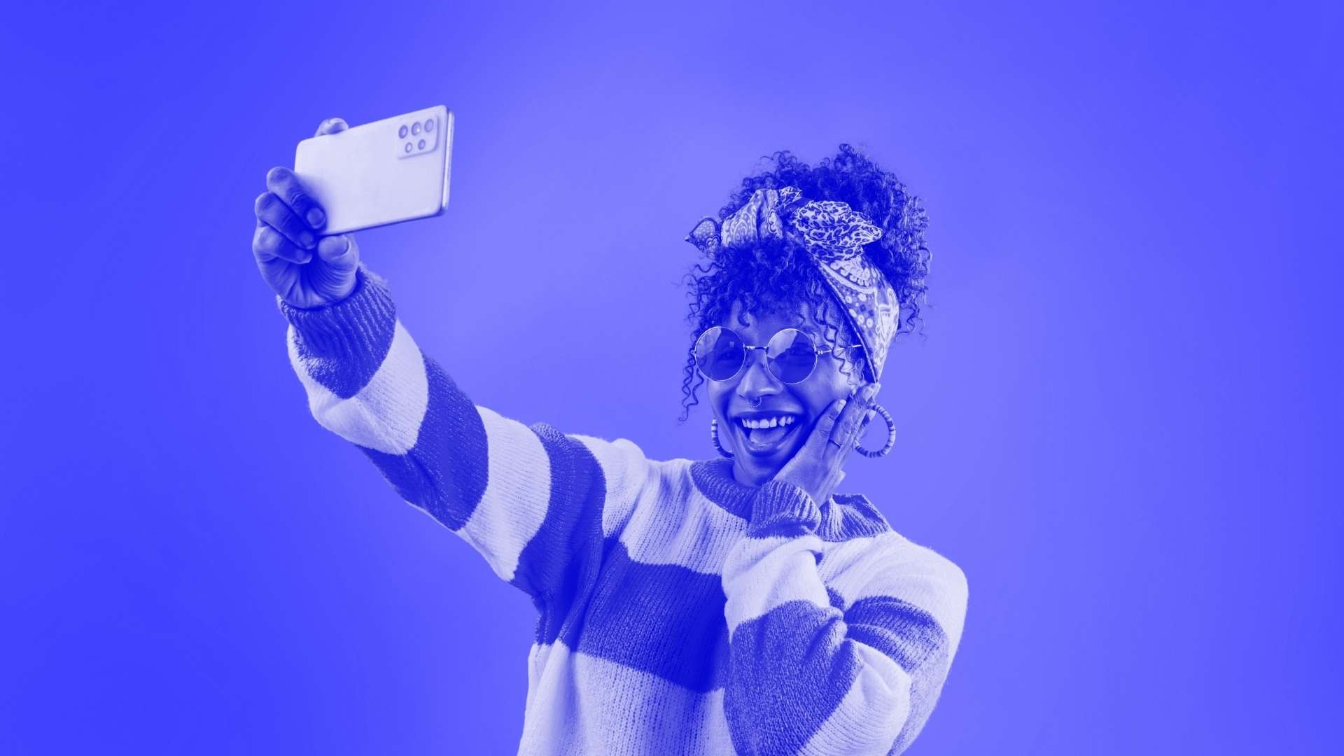A woman posing for a selfie wearing a jumper with thick stripes on it, sunglasses and her hair up in a bun with a bow headband on