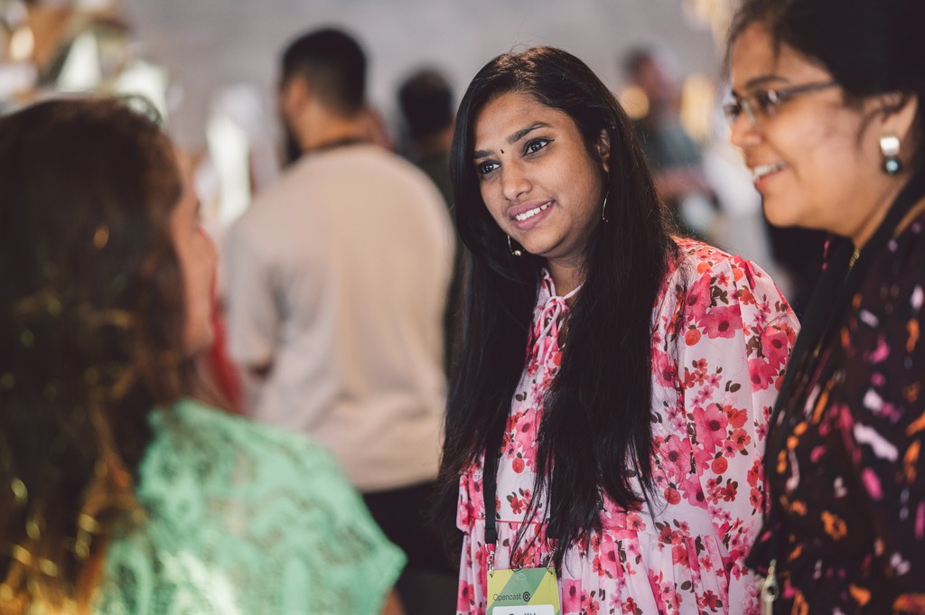 A group of people stand talking in a busy indoor setting. The focus is on an individual wearing a floral patterned outfit, engaged in conversation with others nearby. The background shows several more people gathered and interacting in a softly lit environment.