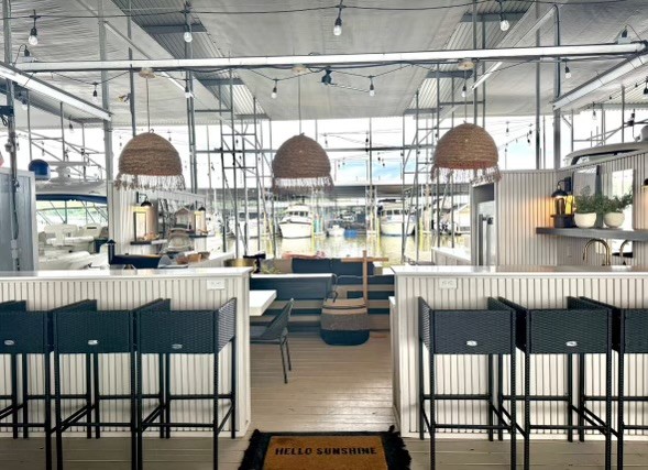 Spacious outdoor dining area overlooking a marina, featuring wicker pendant lights, modern bar stools, and nautical decor, creating a serene ambiance under hanging string lights.