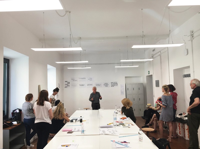 A photo of some students standing in a room while listening to the teacher talking about their letter drawings.