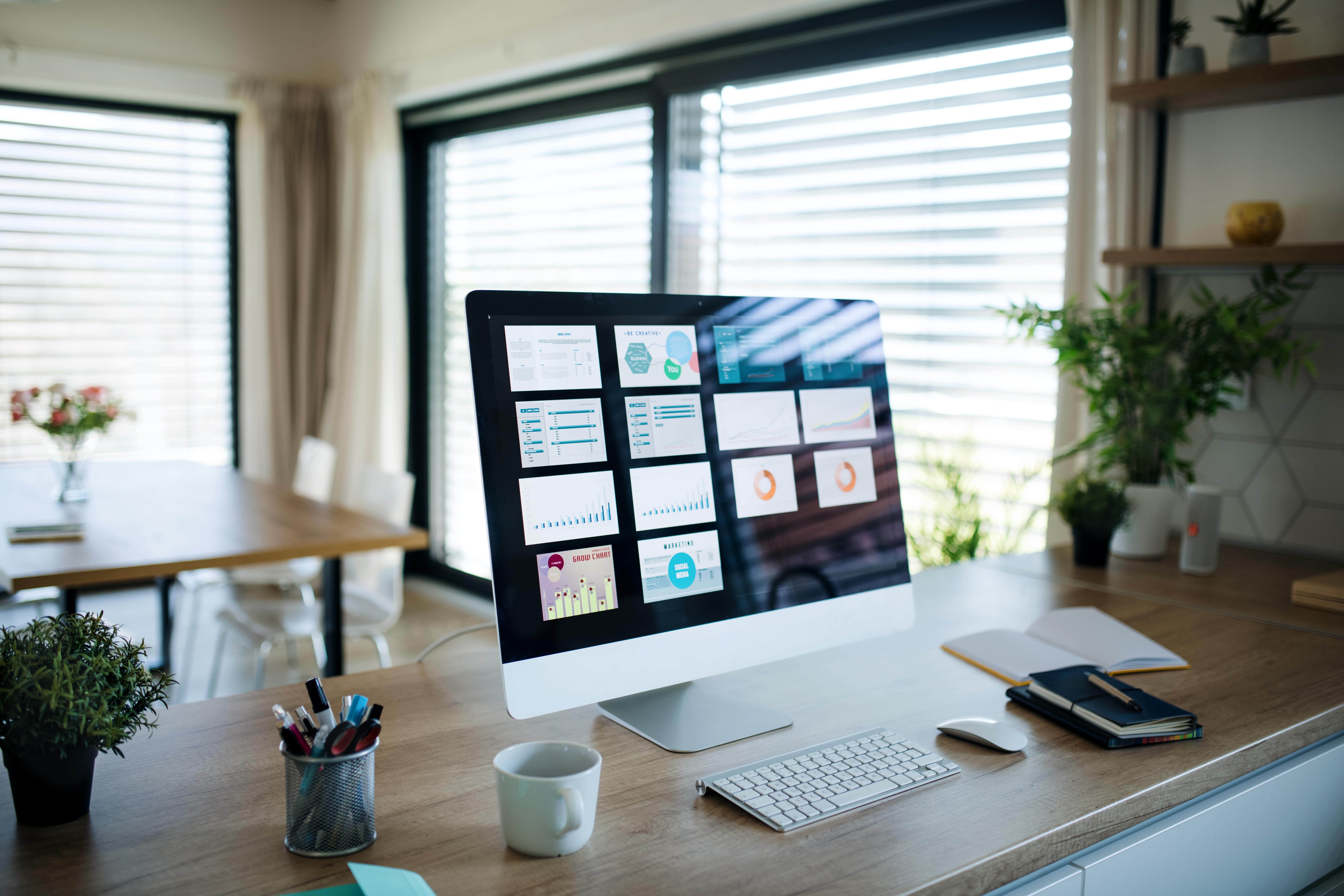A computer monitor displaying a data analytics and marketing dashboard sits on a wooden desk in a modern, well-lit home office. The work-from-home setup includes a sleek keyboard, mouse, coffee mug, and notebooks, with a bright, open-plan living and dining area visible in the background, creating a productive remote work environment.