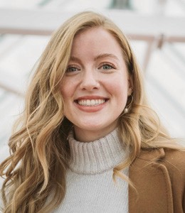 A smiling woman with long wavy hair, wearing a light sweater and a brown coat, stands in a bright indoor setting.