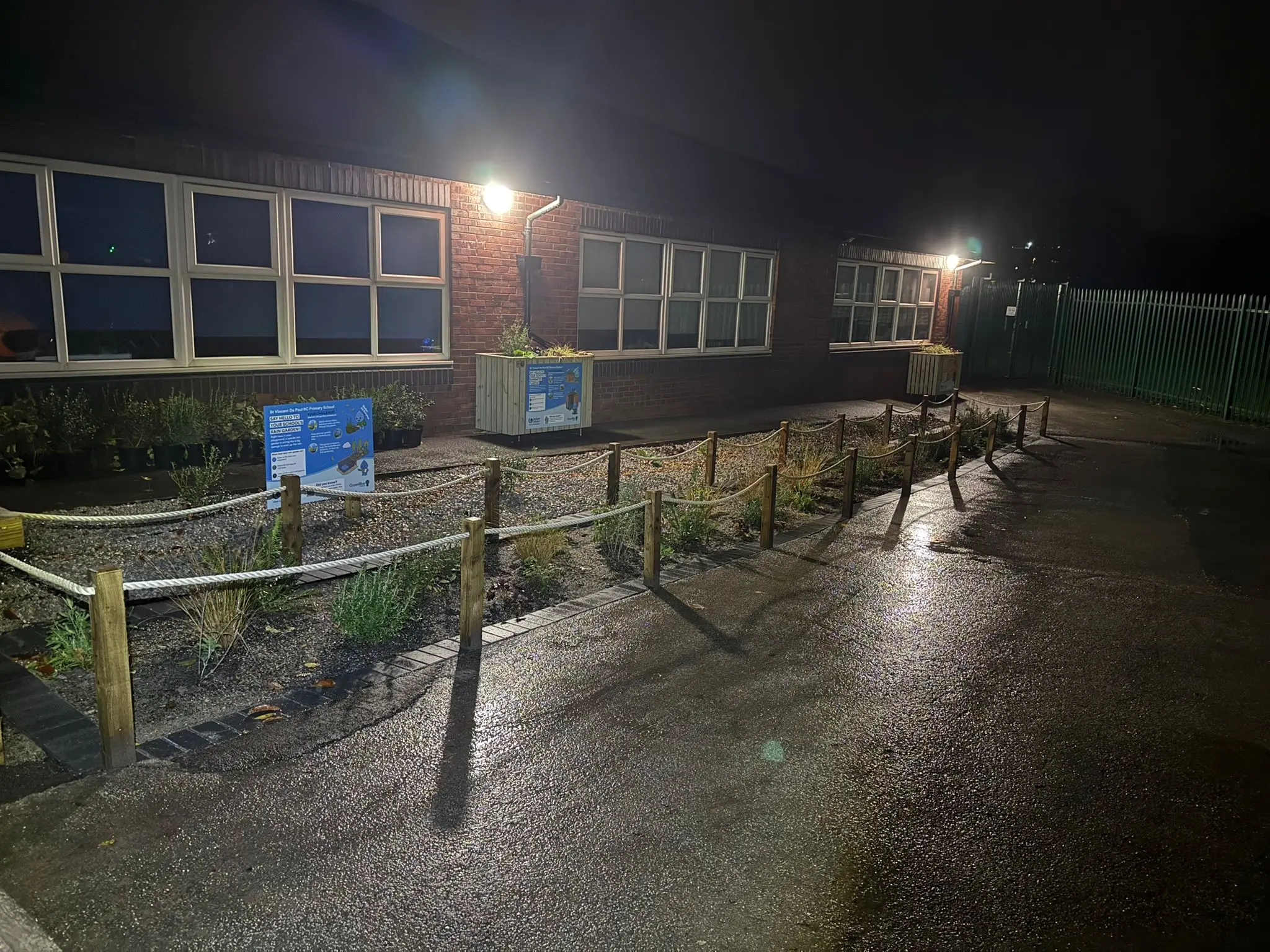 Dimly lit pathway at night, lined with bushes and buildings, with street lamps illuminating the scene.