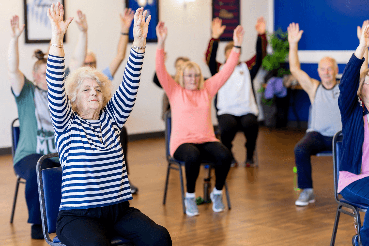 Older adults in a community hall seated in a circle with their arms raised, following a chair exercise class.