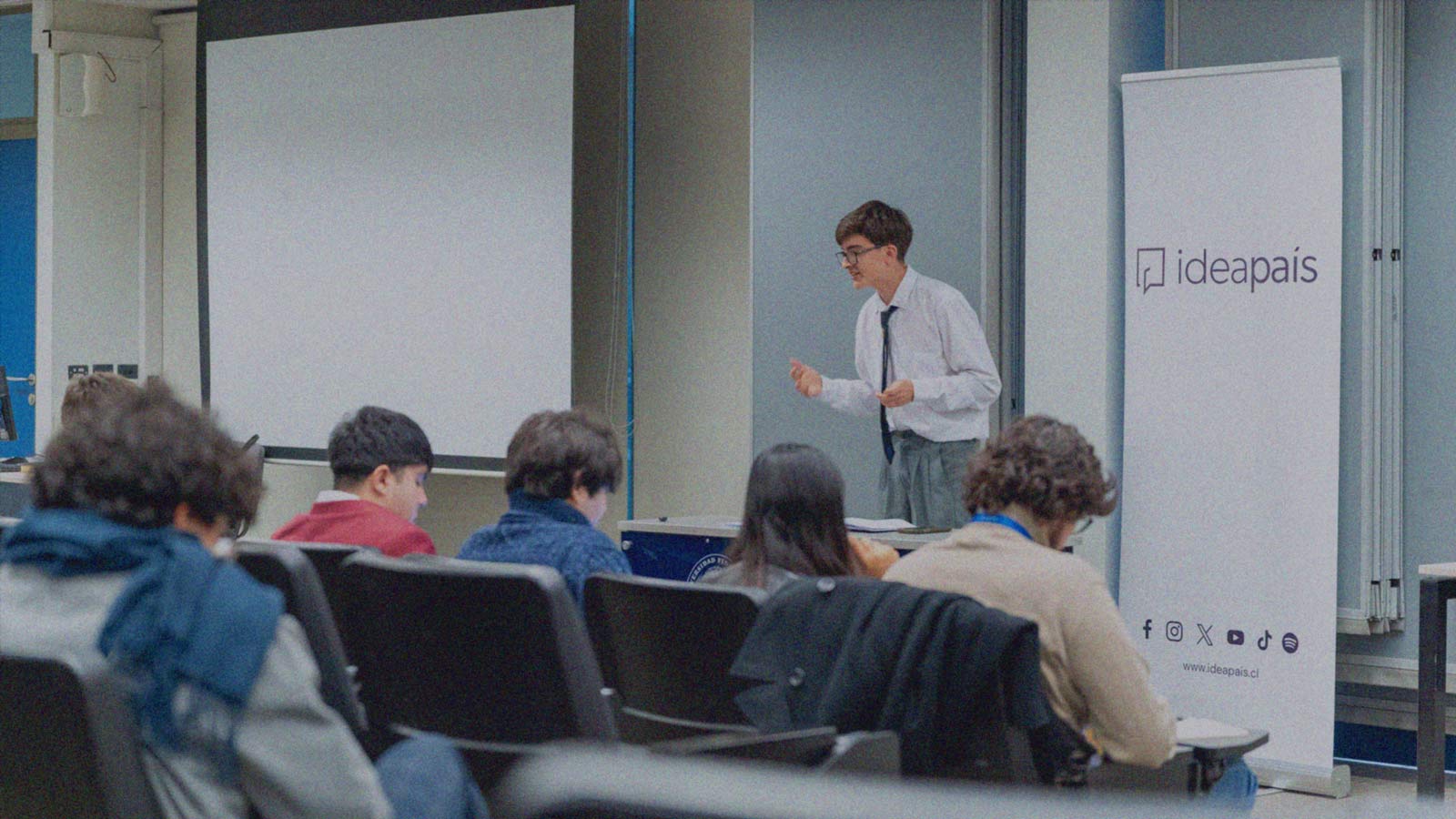 Portada de TIDEIP, programa de formación de IdeaPaís, mostrando una sala en que un escolar expone delante de otros compañeros que le escuchan sentados. 