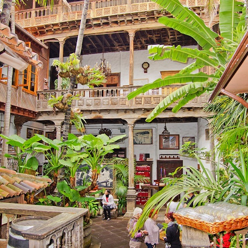 Lush courtyard with people, surrounded by plants and multi-level wooden balconies featuring detailed architecture.