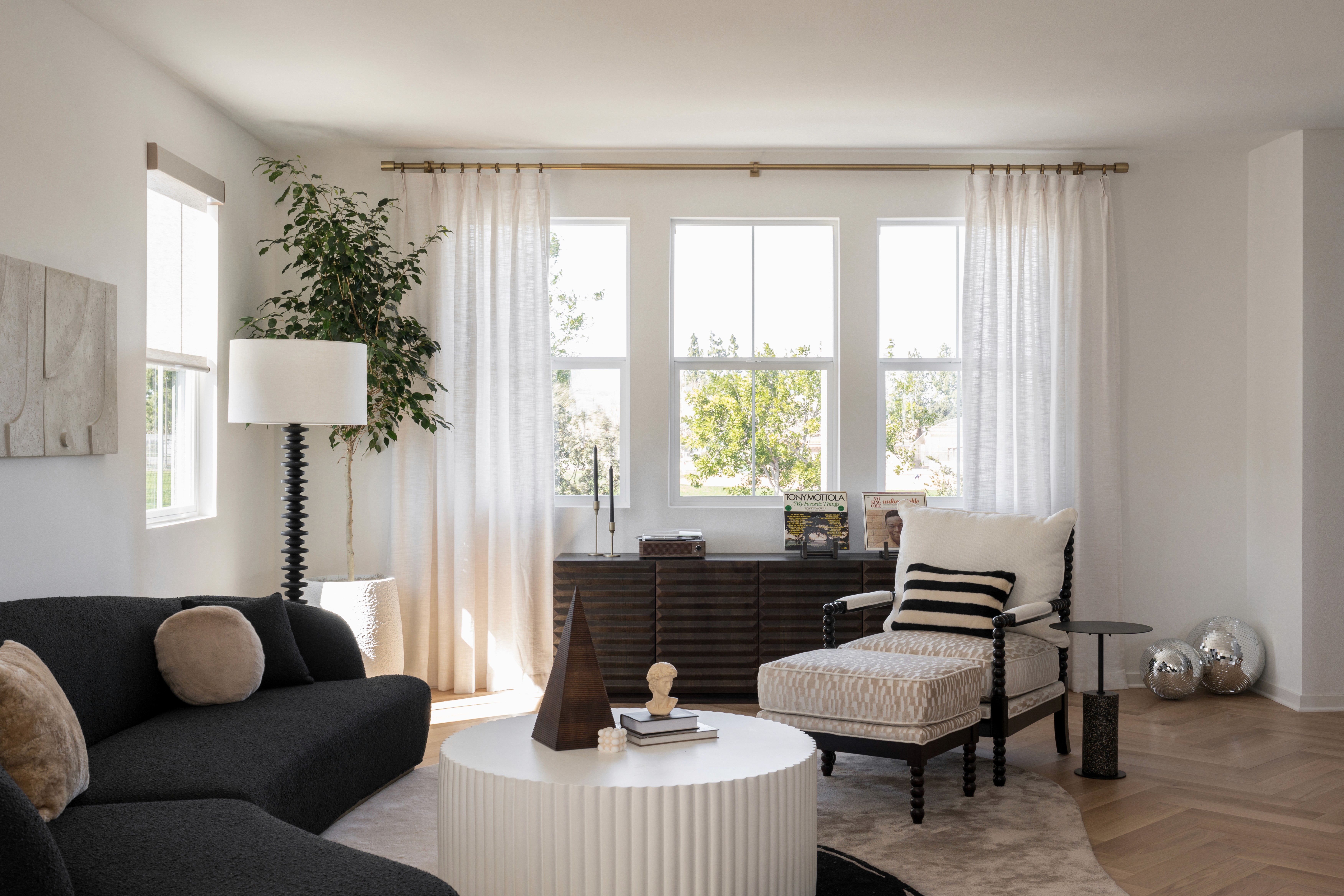 Black kidney bean couch, round, fluted coffee table, large area rug with geometric pattern, modern floor lamp; dark wood console table in background under large windows with floor to ceiling drapes