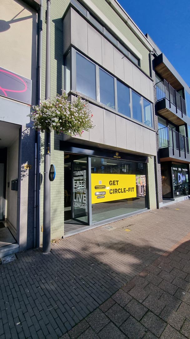 Storefront of Circle Fit Kalmthout with large yellow banner, windows, and clear blue sky; adjacent buildings line the street.