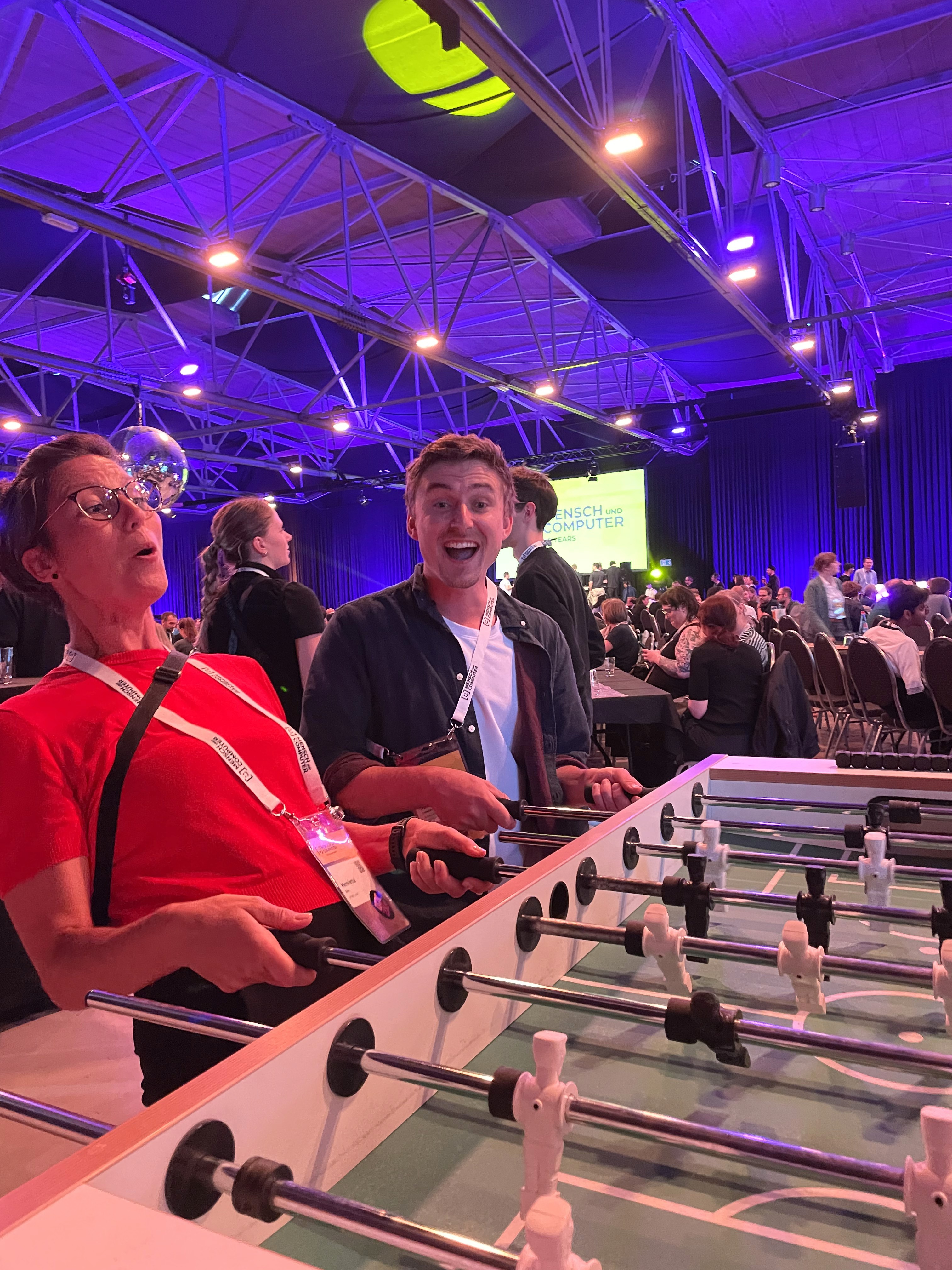 Mehrere Personen spielen Tischfußball in einer großen Halle mit violetter Beleuchtung und sichtbarer Deckenstruktur