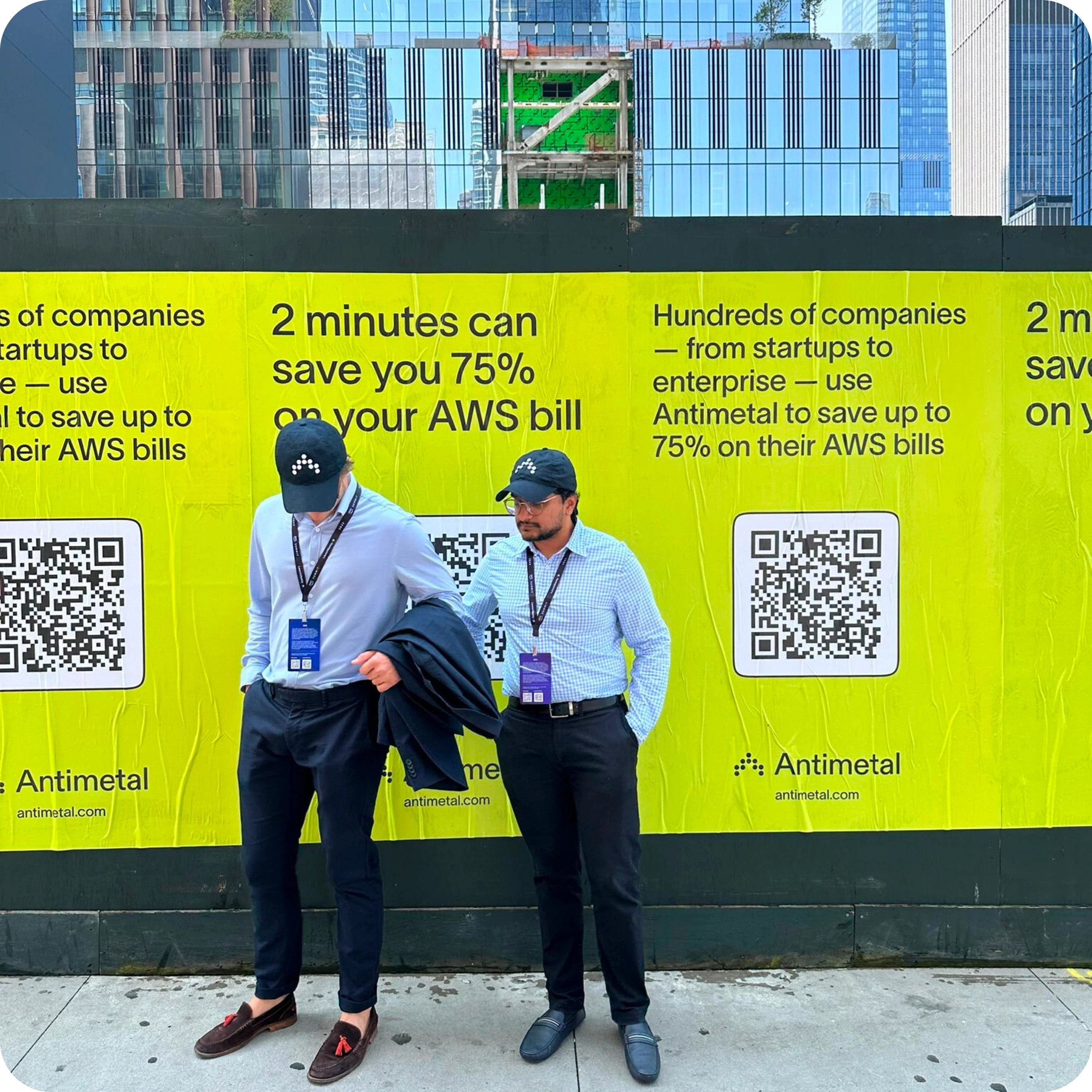 Two men wearing Antimetal caps and lanyards stand in front of a bright yellow advertisement for Antimetal. The ad promotes saving up to 75% on AWS bills in just 2 minutes and features QR codes for more information. The background includes text about Antimetal’s services and is set against a cityscape with tall buildings.