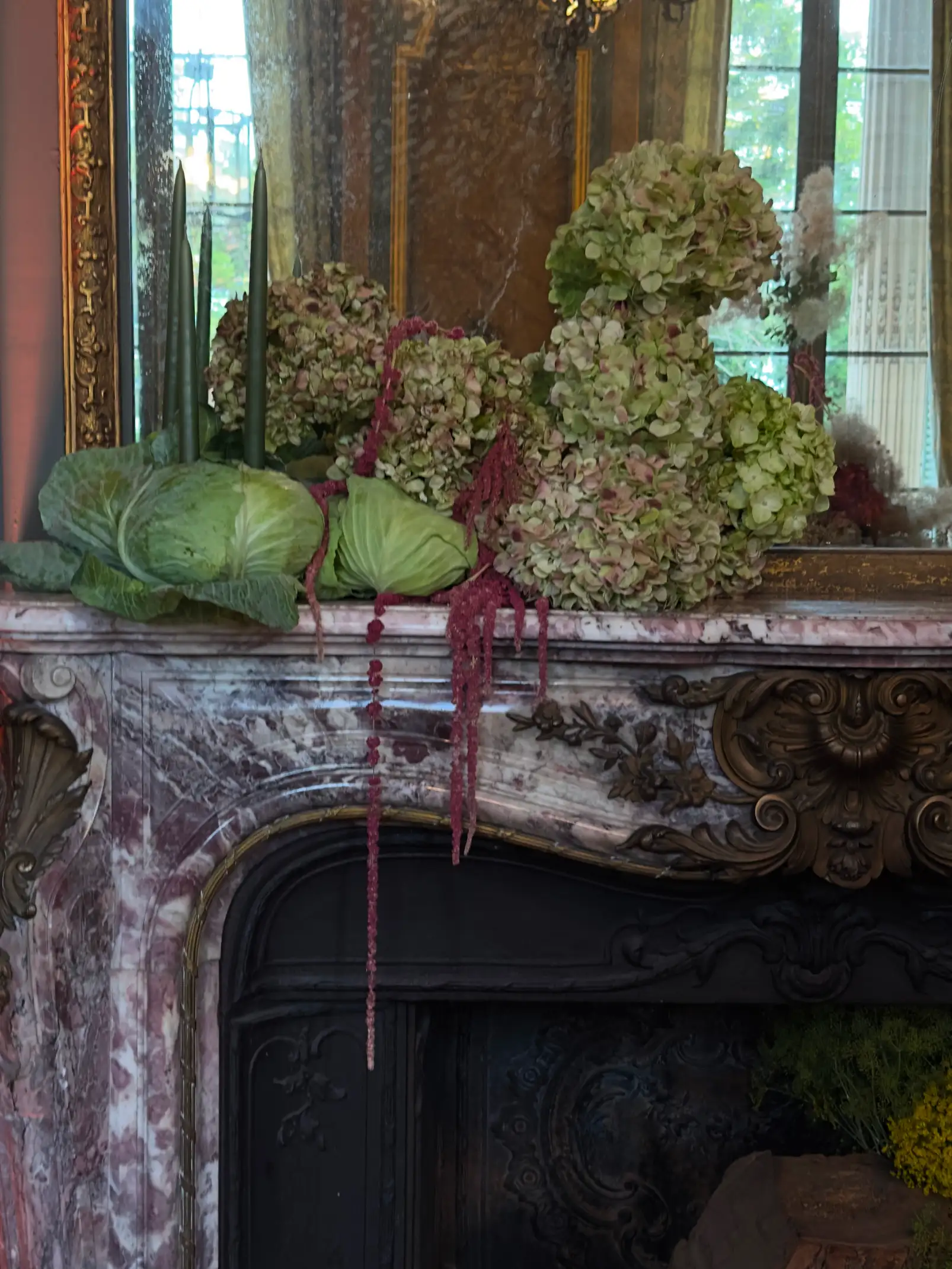 Arreglo floral con hortensias verdes, repollos y amaranto rojo colgante sobre una chimenea de mármol ornamentada, con un espejo dorado de fondo
