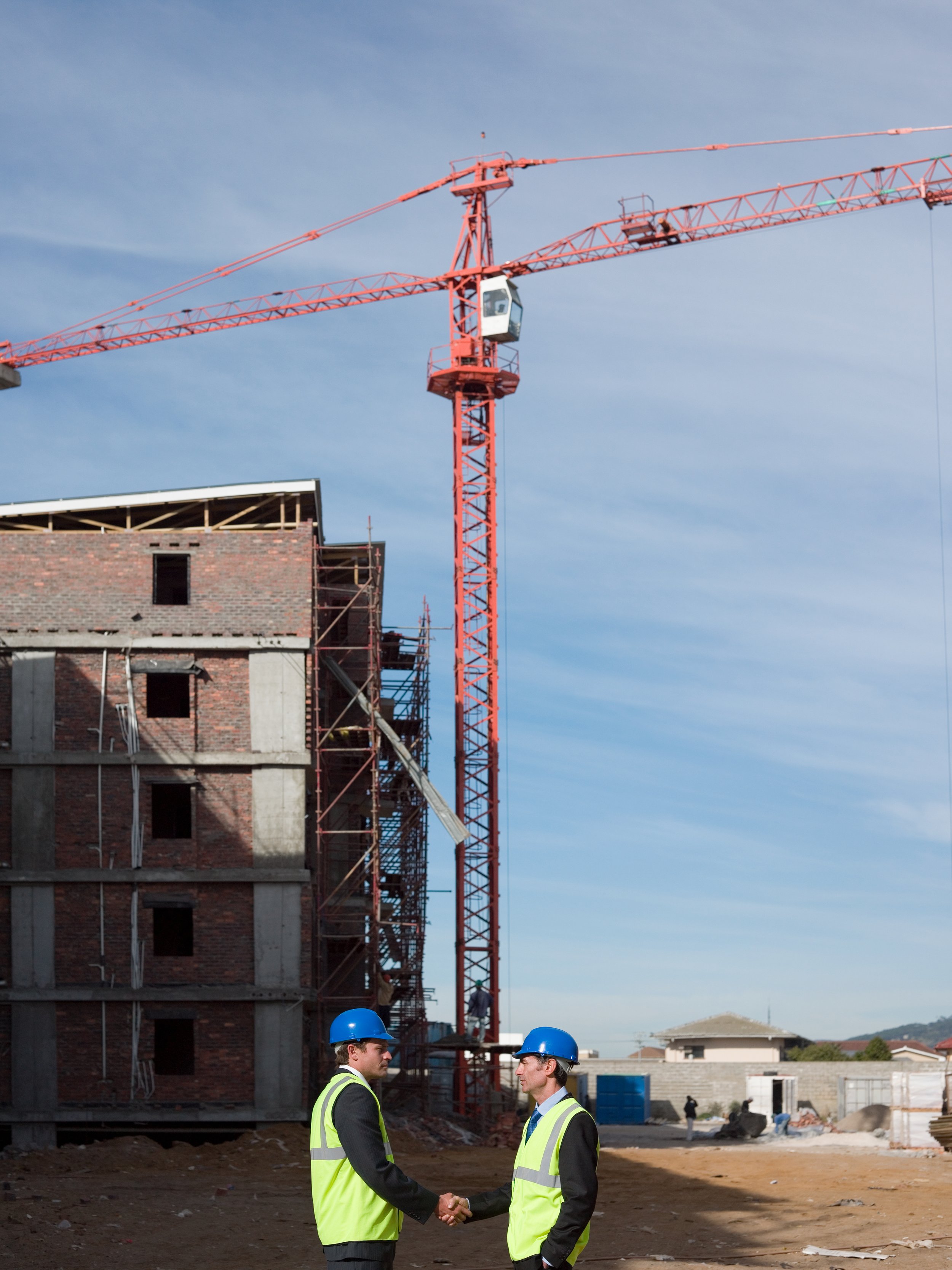 Two people in hard hats and high-visibility clothing at a construction site with a crane and an unfinished building.