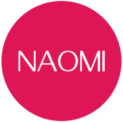 Young person with wavy hair poses confidently in sunlight against a modern building backdrop. Wears a black blazer and silver necklace, conveying elegance.