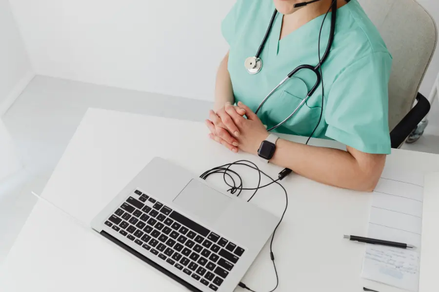 AI nurse communication in action as a healthcare professional in green scrubs sits at a laptop with a stethoscope around her neck and clasped hands resting on the desk.