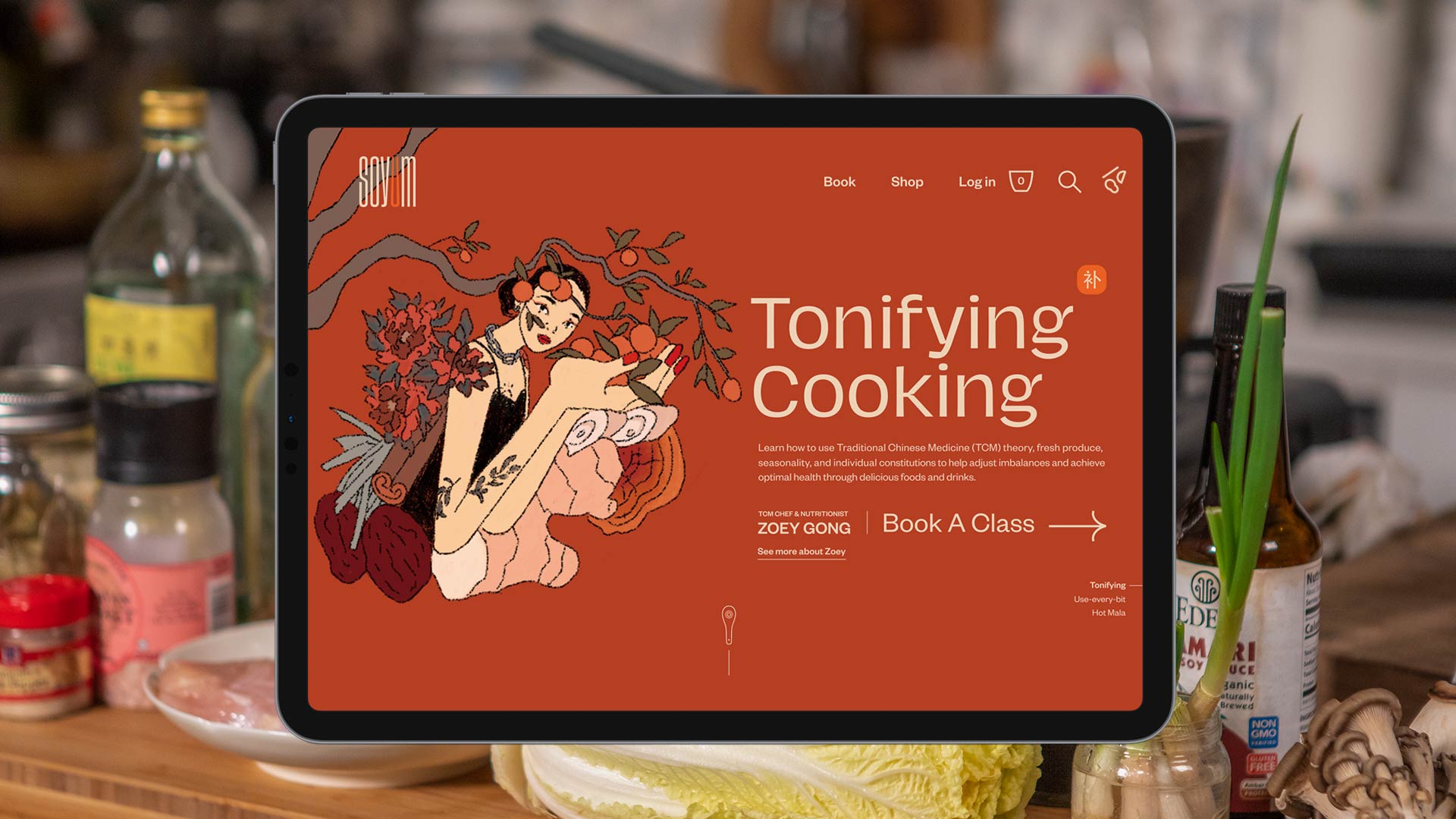 A tablet displaying the website “Tonifying Cooking” is shown. The website features a red background with a stylized illustration of a woman in a floral dress holding oranges, surrounded by Chinese characters and text. The navigation bar includes links for “Book,” “Shop,” “Login,” and “About.” Below the illustration, text reads “Learn how to use traditional Chinese Medicine (TCM) theory, fresh, produce, seasonality, and individual constitution-based foods and drink.” Further down, “Tom Chiu’s Nutritionist” and “Book A Class” are listed with navigation arrows. The tablet rests on a kitchen counter with bottles, jars, and a bowl of vegetables in the background.