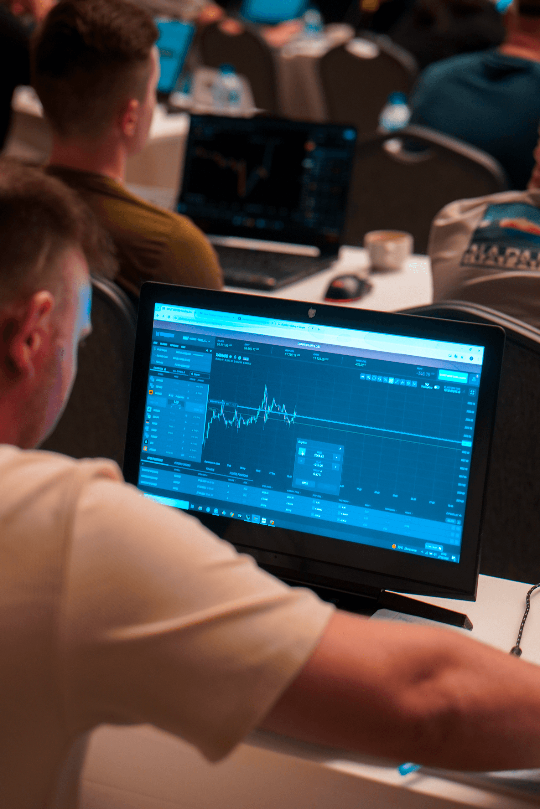 Trzy młode osoby w bibliotece, uśmiechnięte i zgromadzone wokół laptopa. Jedna z nich trzyma podkładkę z notatkami, na stole leżą otwarte książki.