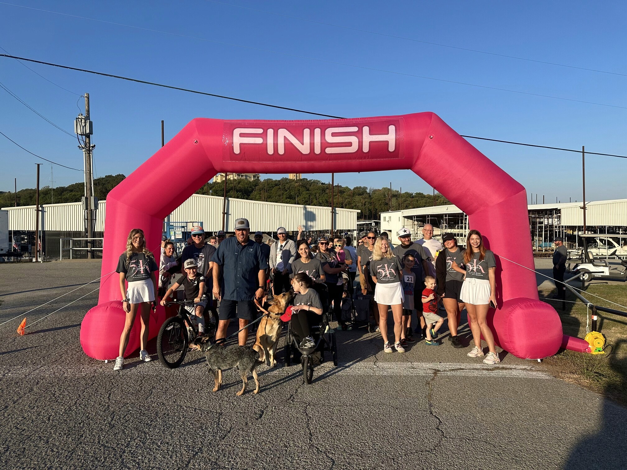 A group of people, some with dogs, gather at a bright pink finish line arch during a community 5K event under a clear blue sky.