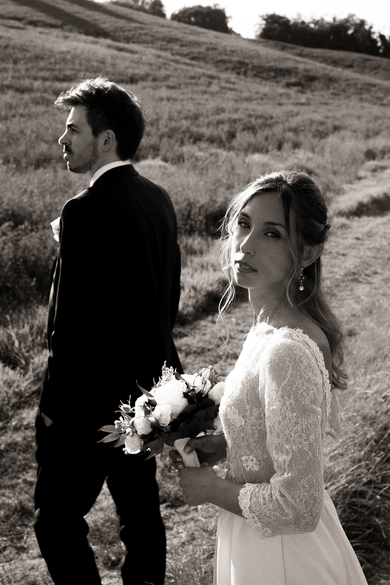 Sposi in bianco e nero nei campi di Villa Peroni Riva, lei con bouquet, lui di profilo, colline sullo sfondo