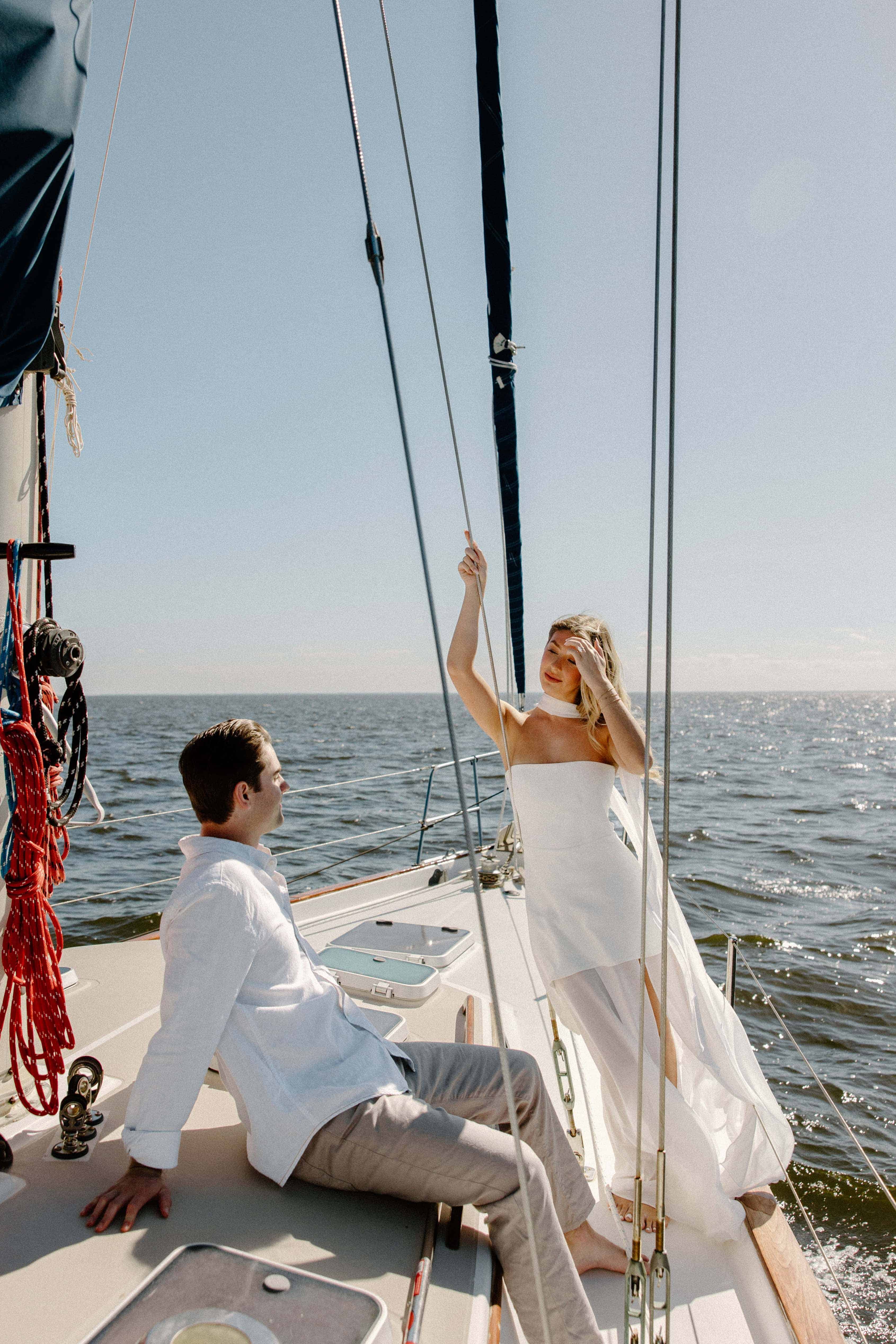A man and a woman are sitting on a sailboat on a body of water with a clear sky. The man is in a white shirt and the woman is in a long, white dress.