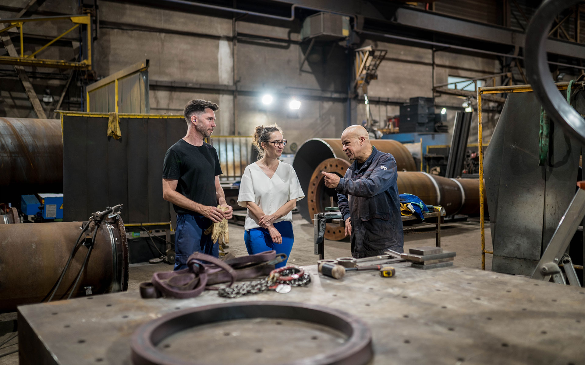 Dans un atelier industriel, trois personnes sont en pleine discussion devant un grand établi métallique au premier plan. Un homme vêtu d'un t-shirt noir et un pantalon de travail, une femme portant un haut blanc, et un homme plus âgé en bleu de travail échangent des idées, ce dernier faisant un geste explicatif de la main. L'arrière-plan révèle un environnement de fabrication avec des pièces métalliques imposantes et des équipements industriels, soulignant une atmosphère de collaboration technique.