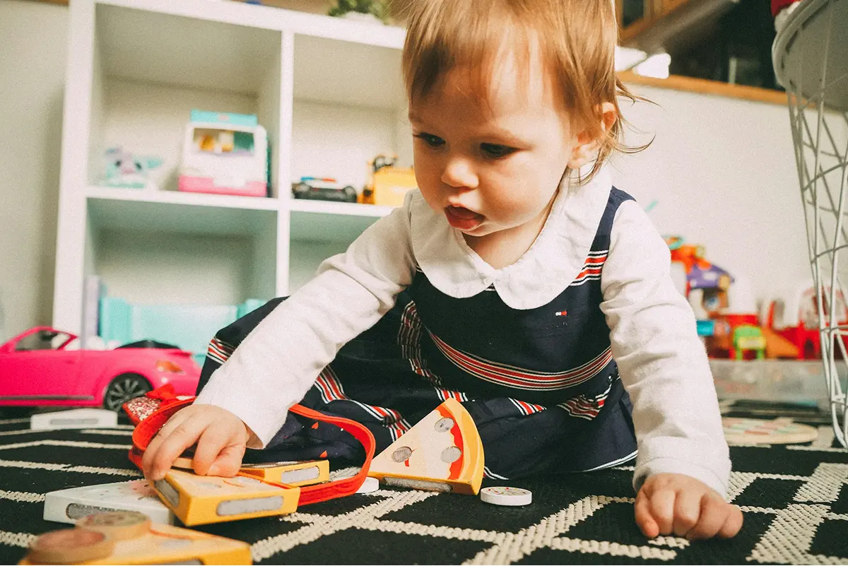 A toddler playing with wooden toy food on a play mat in a bright classroom playroom