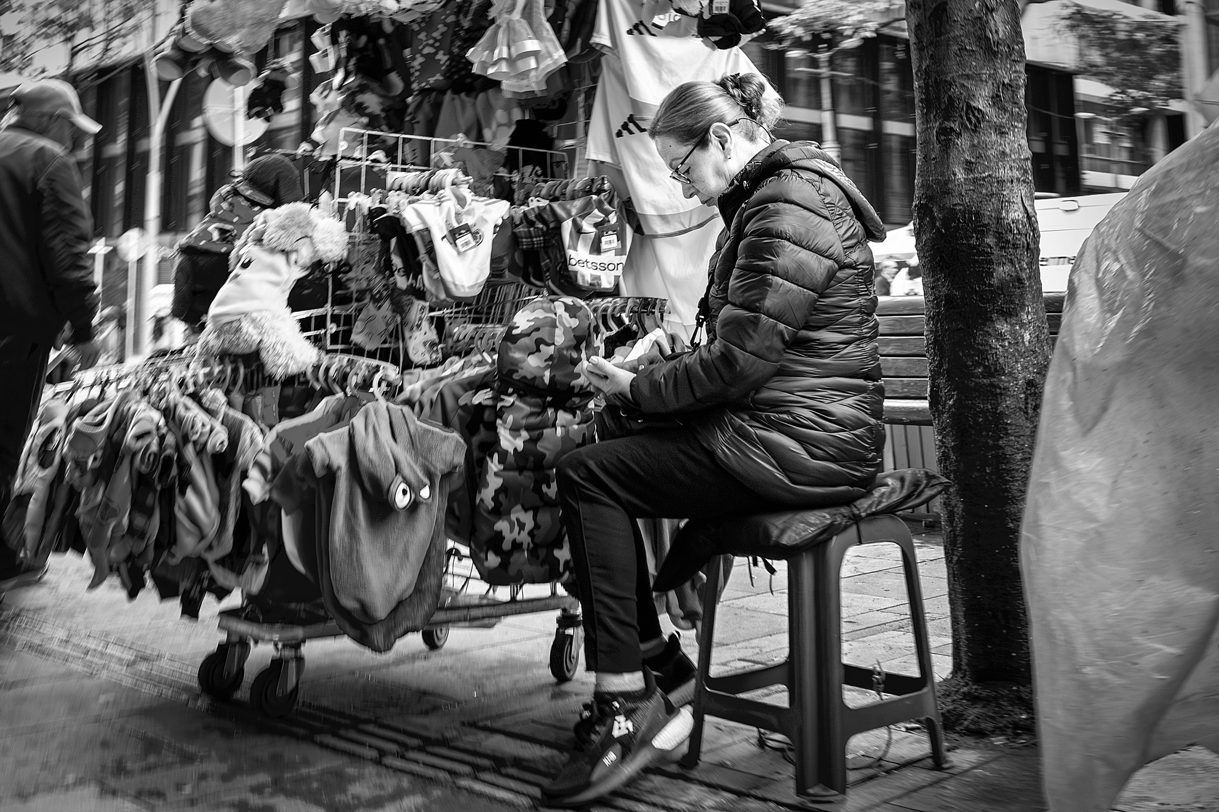 A woman sits on a plastic stool next to a rack of children’s or pet clothing on a busy Bogotá sidewalk, inspecting an item in her hands.