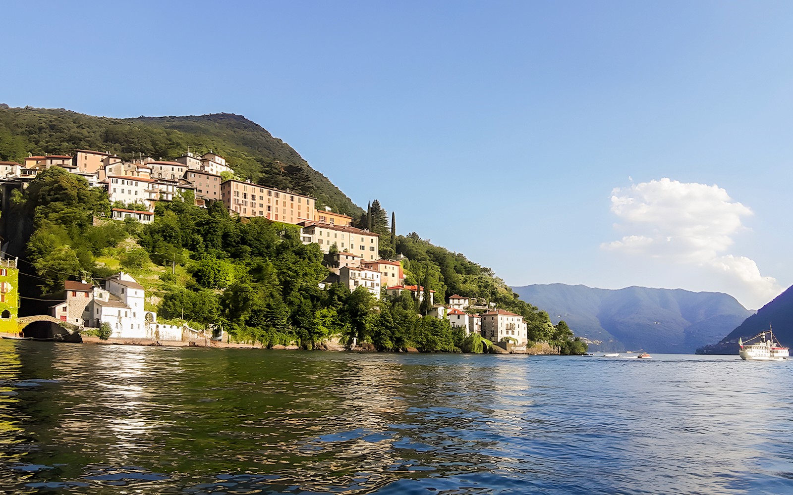 Boat cruising on Lake Como with hillside village and mountains in the background.