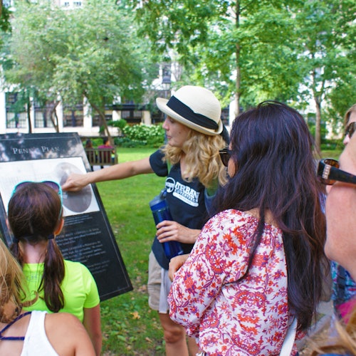 在公园里，一位戴帽子和穿黑色衬衫的女士一边指着信息板，一边对一群人说话。