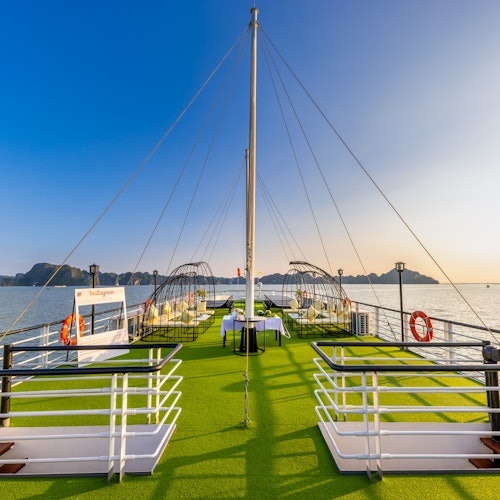 Et båddæk med grøn kunstgræs, borde med stole og redningskranse, der sejler på stille vand under en klar blå himmel ved solnedgang.