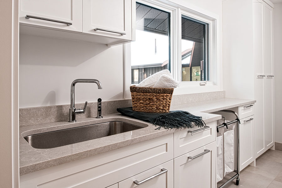 Image of Modern kitchen sink area with white cabinets, grey countertops and laundry sorter by KHD Homes.