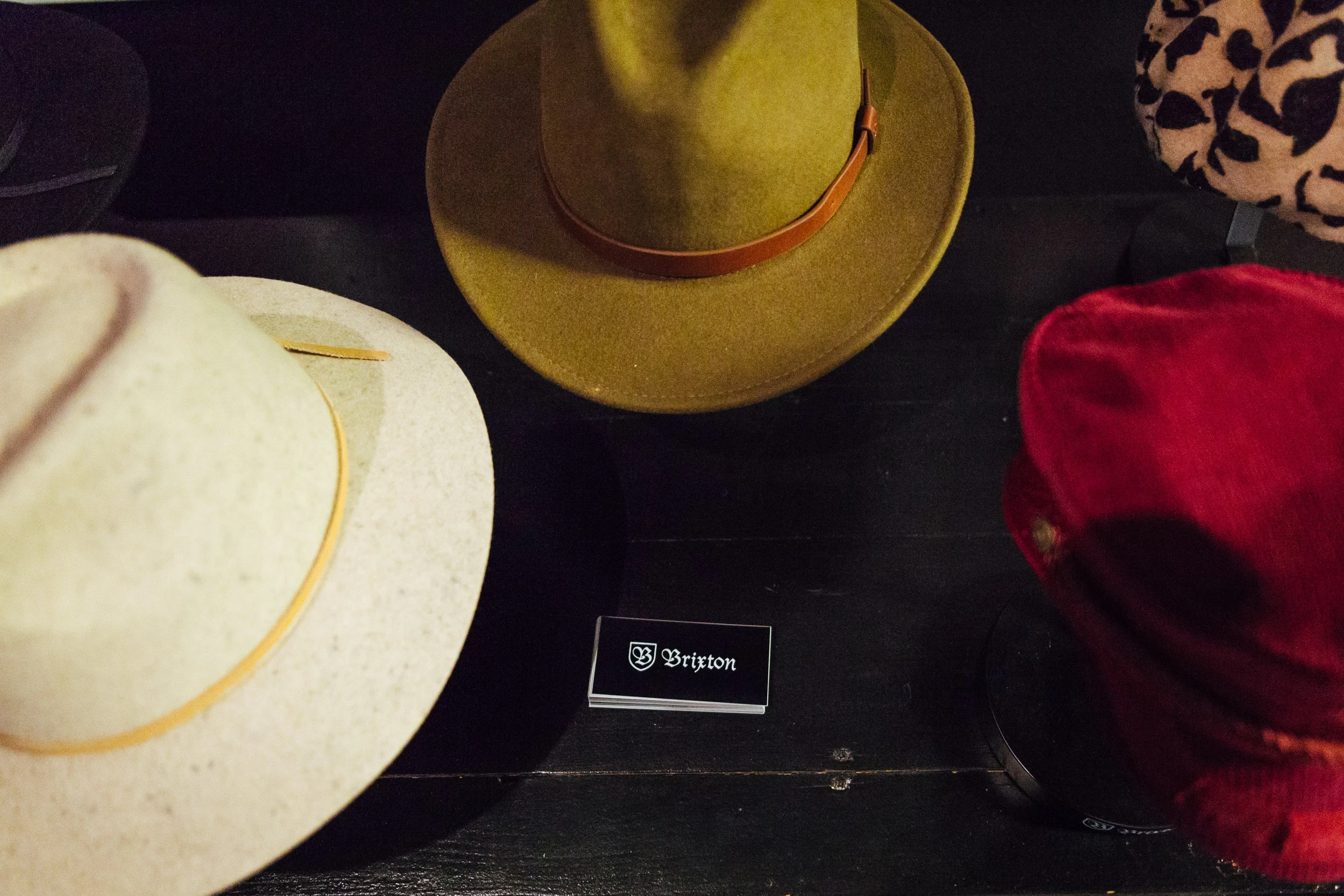 BRIXTON hats in beige, olive and red arranged on a dark table around a small black brand sign.