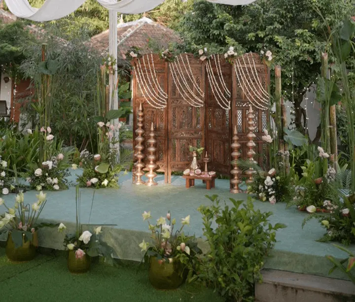 Outdoor wedding mandap at Vedic Village Alleppey with carved teak wooden backdrop, jasmine garland drapes, brass nilavilakku lamps and tropical garden setting