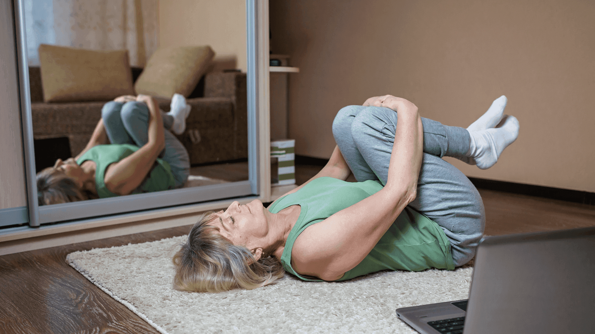 Woman practicing yoga at home, lying on a beige carpet in a knee-to-chest pose (Apanasana) wearing a green tank top and grey pants, with a laptop nearby for following an online class