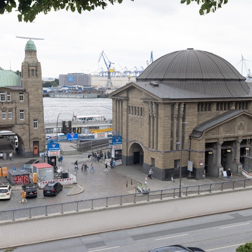 An urban scene featuring a large stone building by the waterfront, with cranes and industrial structures in the background. People and cars are visible.