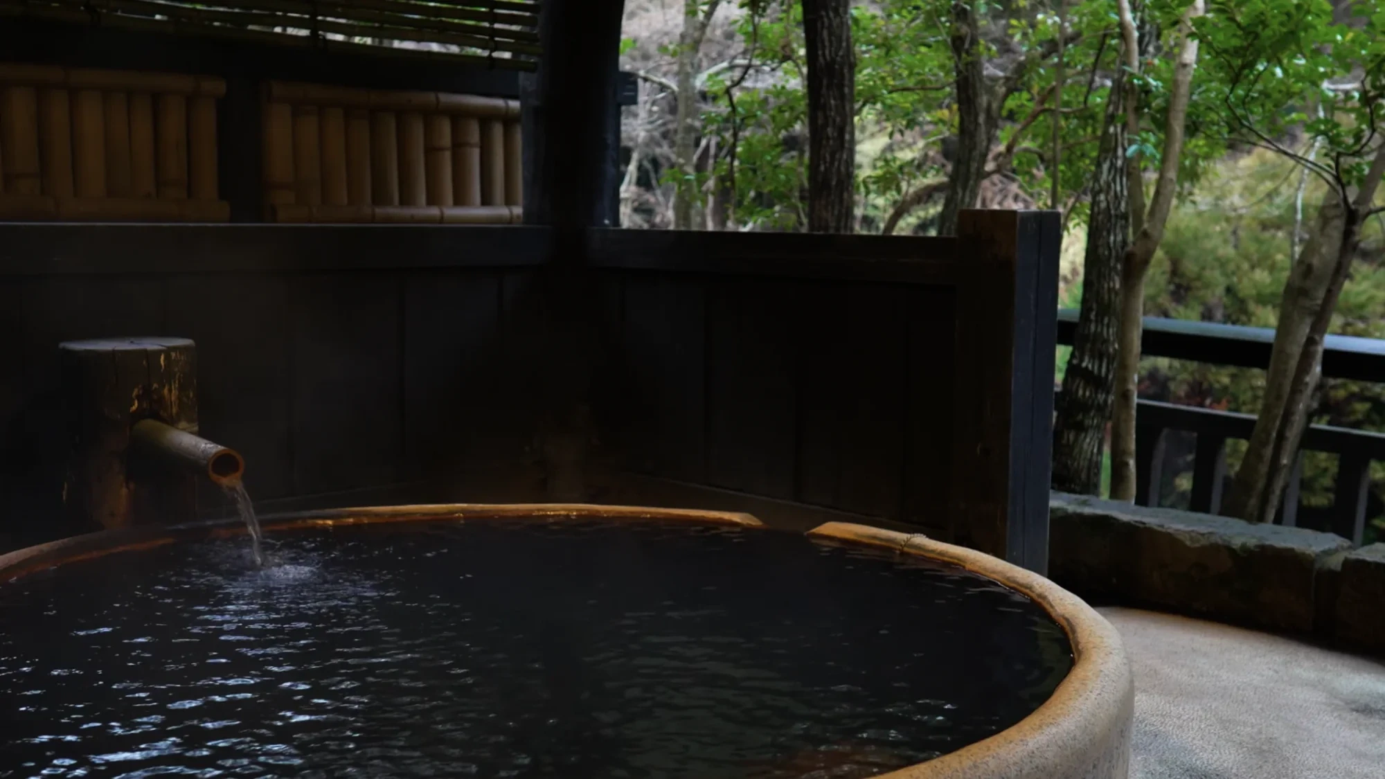 A round bathing tub at Kurokawa Onsen with a spout and a timber rail, forest beyond