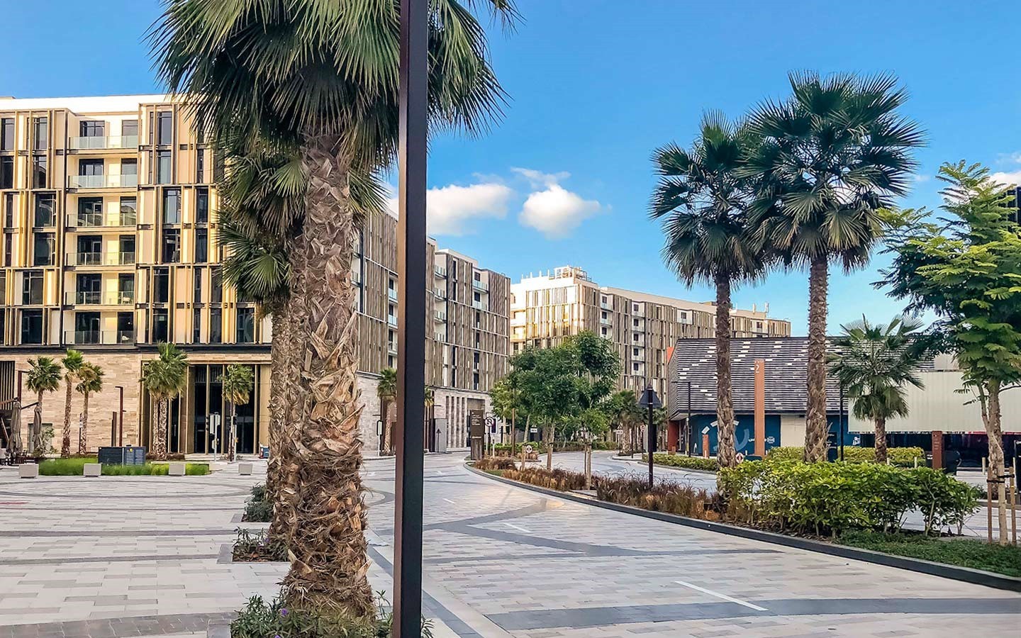 A wide pedestrian walkway and street lined with palm trees and modern residential buildings on Bluewaters Island under a blue sky.