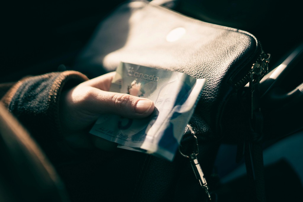 A hand holding a blue Canadian five-dollar bill next to a black leather purse, representing government tax benefits and cash payouts for newcomers.