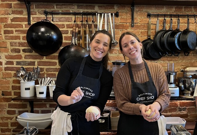 Two women wearing aprons stand in a kitchen with exposed brick walls, holding dough and smiling at the camera—looking like media pros collaborating in an agency environment. Cooking utensils and pots hang on the wall behind them.