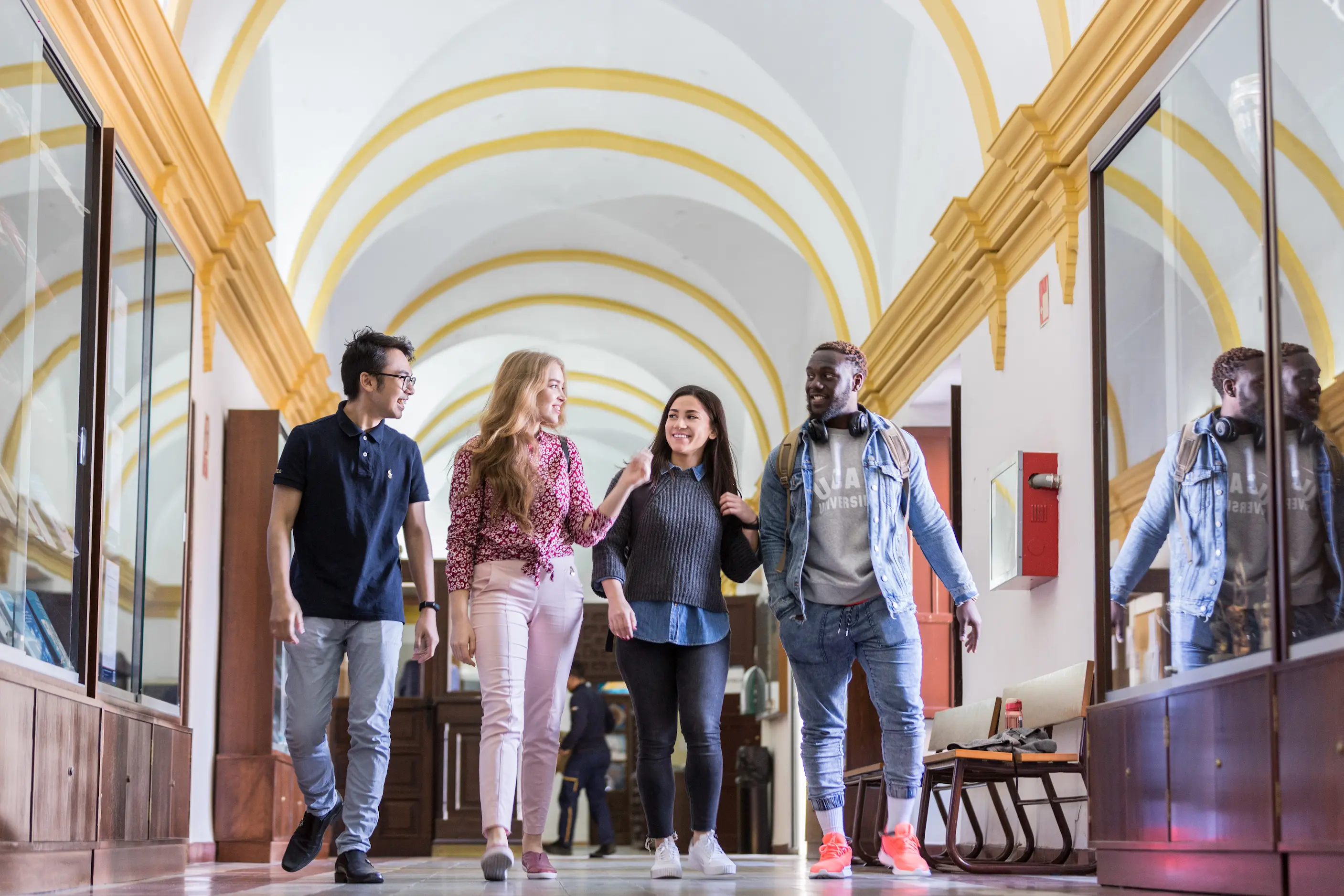 A group of students carrying backpacks walk together through a campus with arched corridors and stone pillars, suggesting an academic setting similar to UCAM campus in Mumbai.