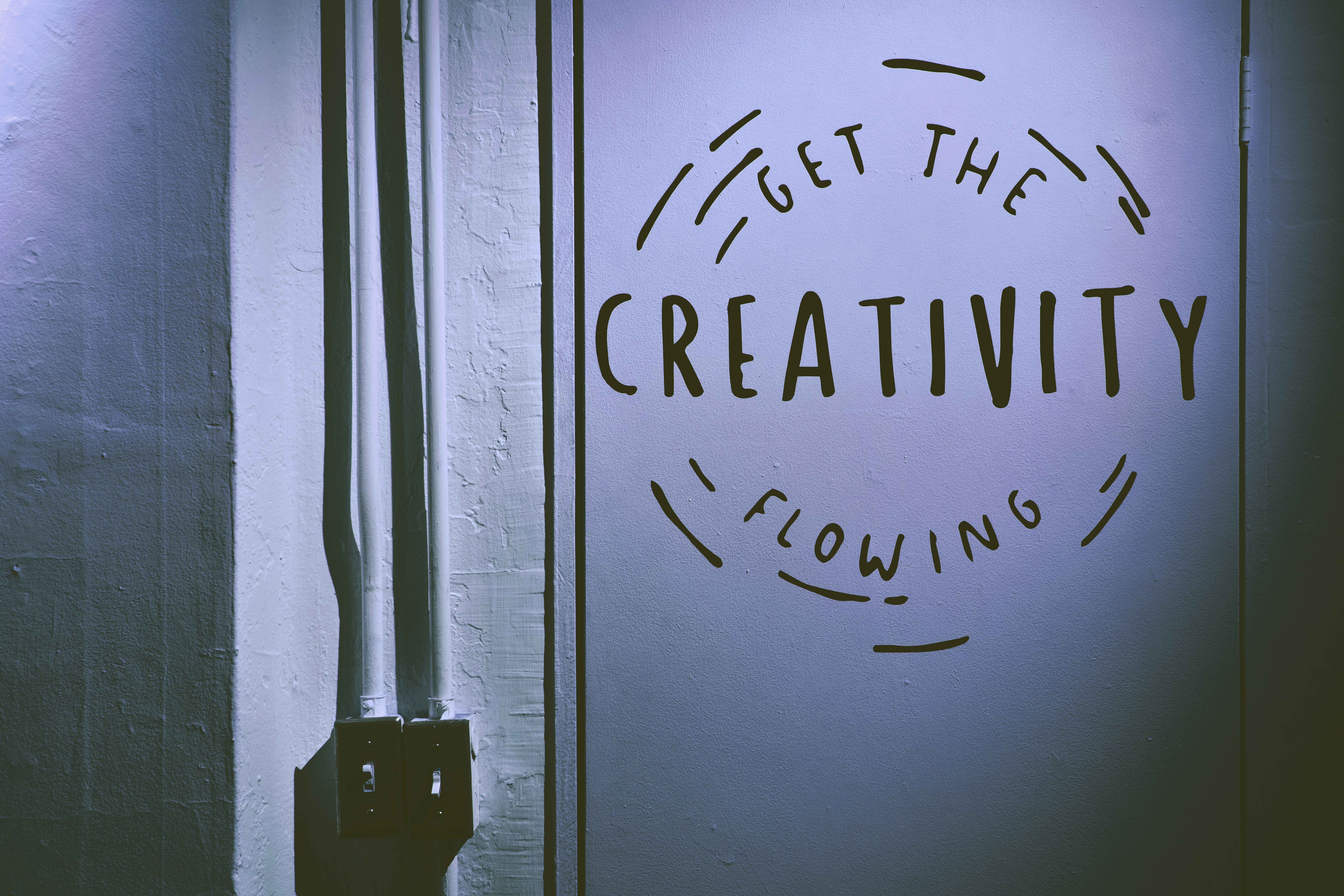 A dimly lit wall features the motivational phrase "Get the Creativity Flowing" in bold black letters, surrounded by simple decorative lines, next to visible electrical conduit, highlighting an industrial, artistic ambiance.