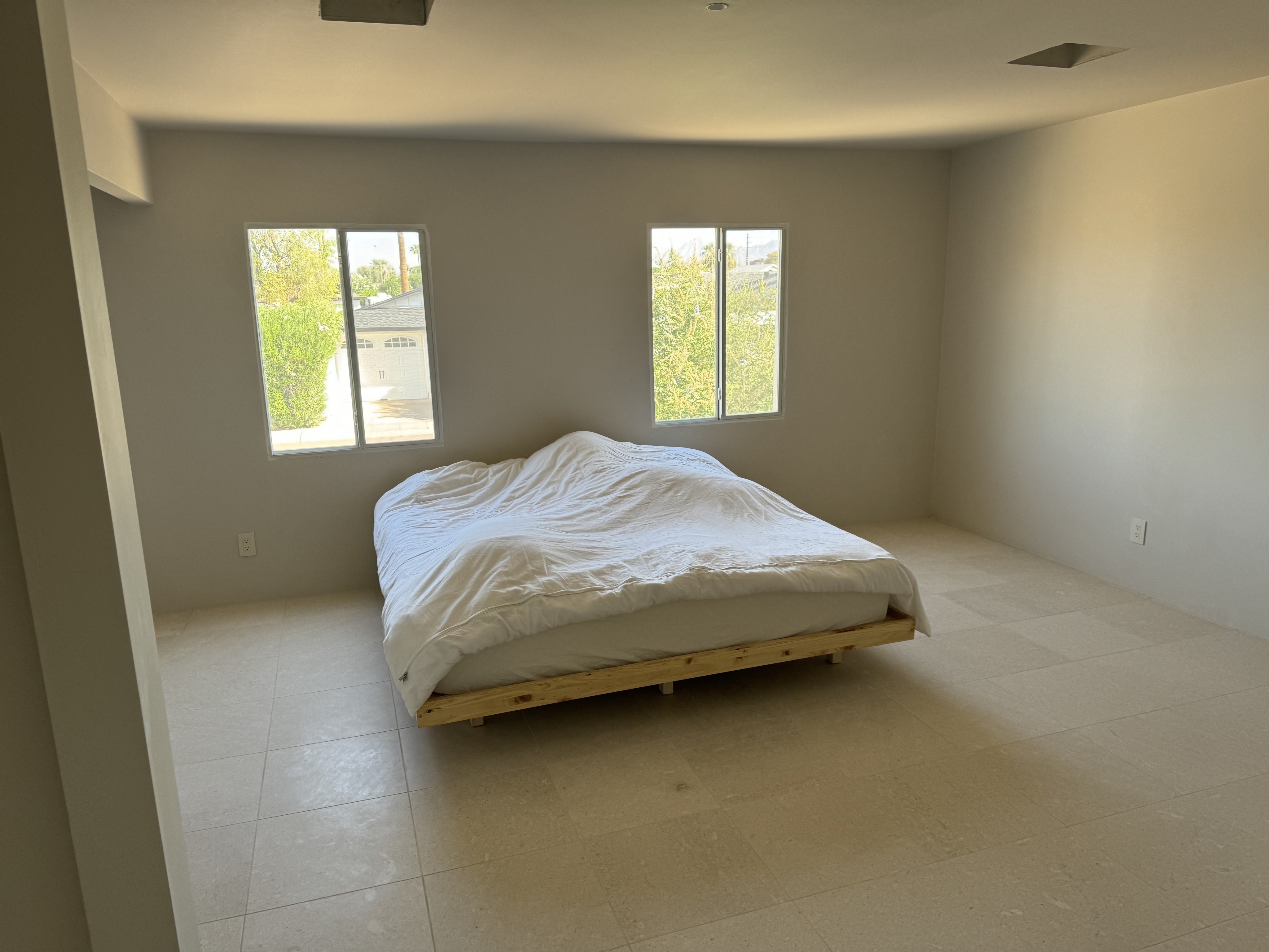 Modern bedroom with a simple bed frame, white bedding, and large windows letting in natural light in a new home under construction.