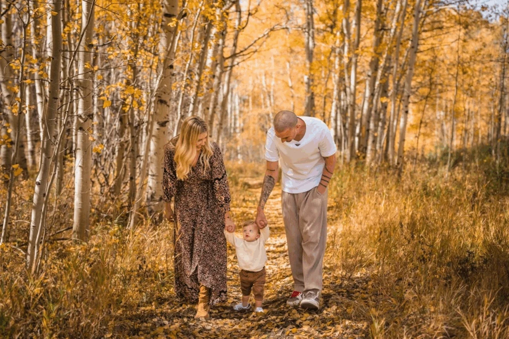 Couple walking through vibrant fall foliage while holding their child's hands, with colorful autumn leaves surrounding them.